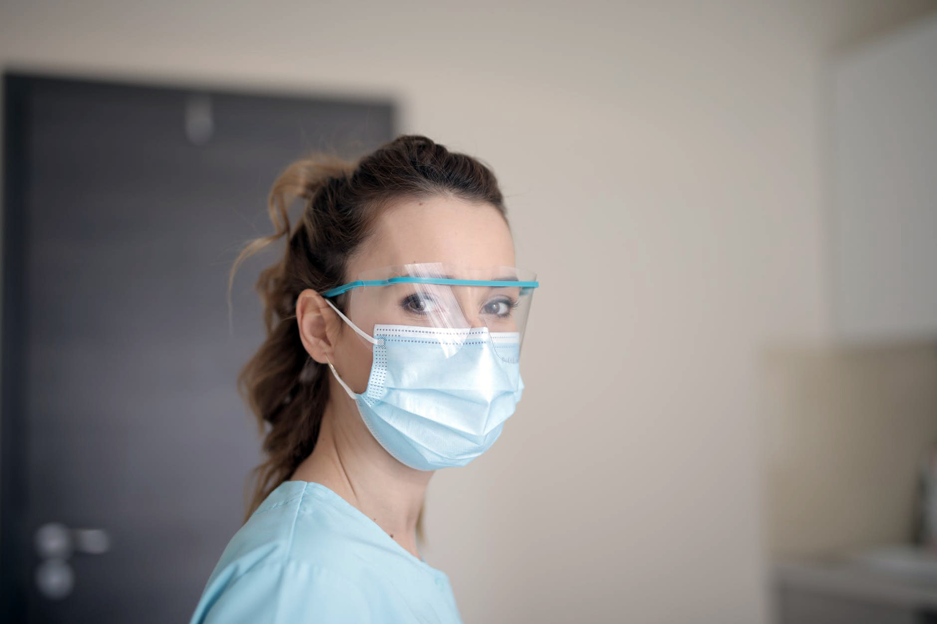 A healthcare worker wearing a blue medical face mask, face shield, and light blue scrubs in an indoor clinical setting.