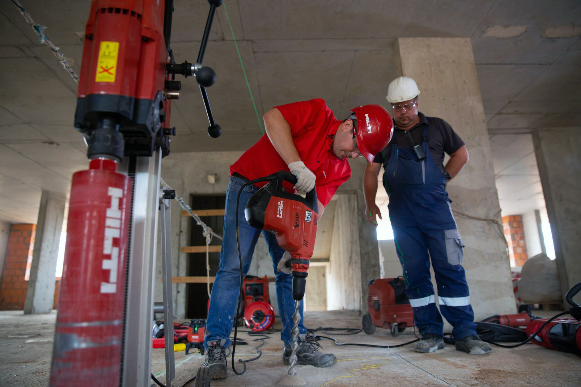 A construction worker in a red helmet operates a heavy-duty drill on a concrete floor, with another worker in blue overalls standing nearby.